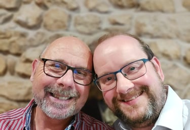 Two smiling men with glasses and beards pose together against a rustic stone wall background.