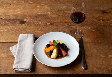 A minimalist, overhead shot of a rustic Brazilian restaurant table. A plate of gourmet food with vibrant colors is centered, surrounded by a linen napkin and a single glass of red wine. Scandinavian aesthetic, warm lighting.