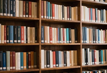 A stack of hardcover books on a white desk in a library with a computer monitor and blurred bookshelves.