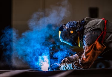 Professional welder in protective gear and mask performing metal fabrication with bright blue sparks and smoke.