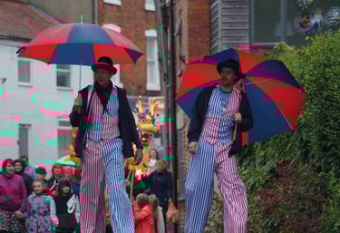 Two circus performers on stilts wearing striped costumes and holding red and blue umbrellas in a street parade.