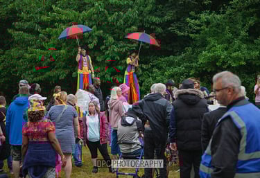 Performers on stilts with colorful umbrellas entertain a diverse crowd at an outdoor community festival.