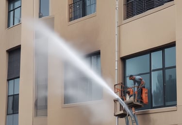 Technician using hot water pressure washer on a commercial building exterior on a sunny day.