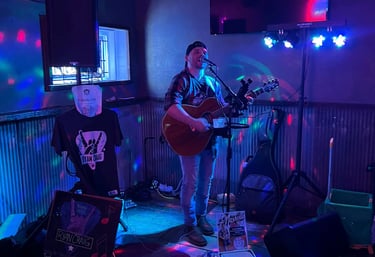 Musician Ryan Craig playing acoustic guitar and singing on a small stage with blue and purple disco lights.