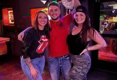 Three smiling friends posing together at a dimly lit bar with colorful neon party lights.