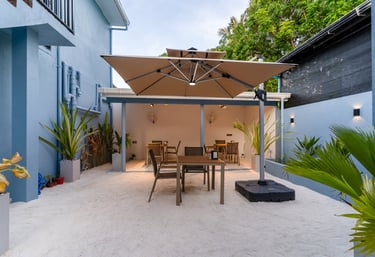 Modern outdoor cafe seating area with a large tan umbrella, dining tables, and tropical plants.