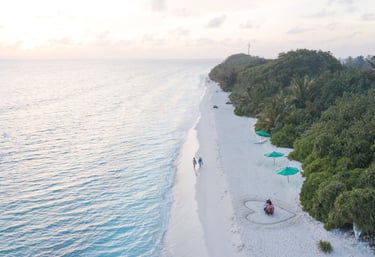Aerial view of a couple walking on a white sand beach at sunset in the Maldives, with lush green trees and turquoise water.