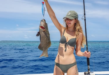 A smiling woman on a boat holds up a large spotted grouper fish caught on a blue lure while deep sea fishing.