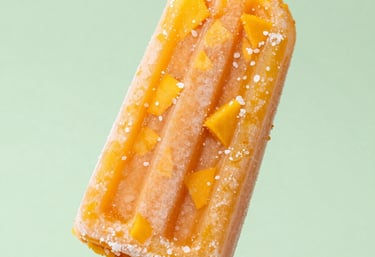 A close-up of a bright orange mango popsicle, with real fruit pieces and a dusting of sea salt, held against a soft light green background. South American summer lighting, crisp and appetizing.