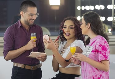 Maquilladora profesional aplica rubor a una mujer sonriente mientras sus amigos disfrutan de bebidas