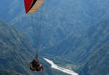 Tandem paragliding over a deep mountain river valley with a red and white paraglider wing.