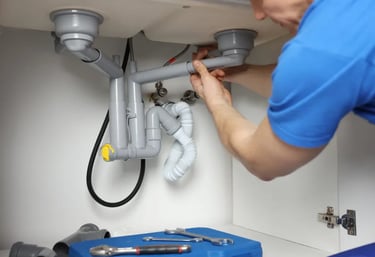 Professional plumber in a blue shirt fixing or installing plastic drainage pipes under a kitchen sin