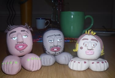 Three handmade clay head sculptures with feet on a wooden table near a green coffee mug.