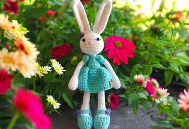 Conejito de peluche blanco hecho a mano con ganchillo, vestido de azul, en un colorido jardín.