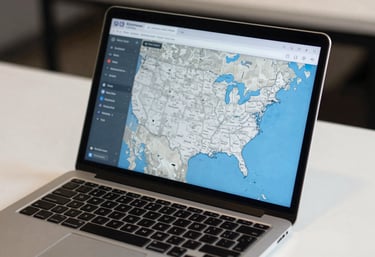 A modern laptop on a white desk displaying a digital map of the United States, with a blue and grey interface, reflecting nationwide business services.