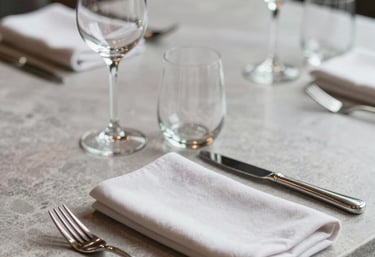 A close-up photograph of a sophisticated restaurant table setting with fine glassware and a cloth napkin on a light grey stone surface. International / Global luxury style.