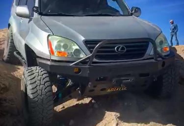 A lifted silver Lexus GX 470 with off-road tires and a steel bumper climbing over rocky desert terrain.