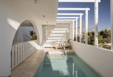 Slim lap pool with pale blue water and beige stone tiling framed by white arches and pergola beams.
