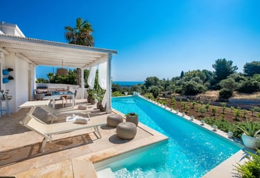 Long Mediterranean pool with bright turquoise water and cream stone tiling overlooking a coastal lan