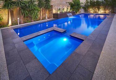 Modern rectangular pool with integrated spa, deep blue illuminated water and dark grey stone tiling.