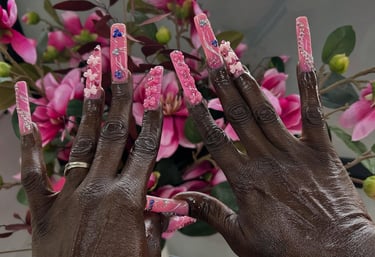 Extra long pink acrylic nails with 3D floral art and glitter accents in front of flowers.
