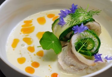 Elegantly plated fish dish with courgette, blue edible flowers, dill, and saffron oil in a white bowl