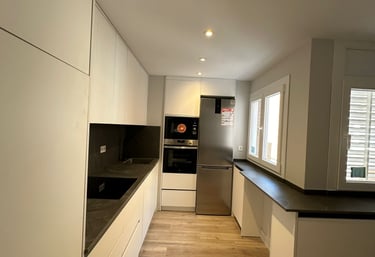 Cocina alargada, muebles blancos, encimeras oscuras, suelo madera.