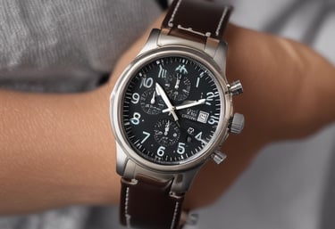 A silver luxury wristwatch with a dark, detailed dial featuring multiple sub-dials and a metal bracelet is placed on a wooden surface. The watch has a polished bezel and luminous hour markers, and the watch hands are prominently visible.