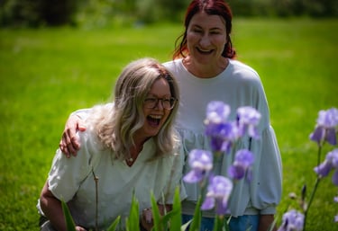 Two laughing women in white tops enjoying blooming purple iris flowers in a sunny green garden.