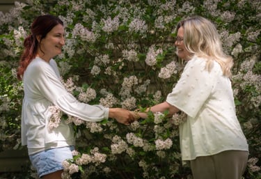 Two women shaking hands outdoors in front of a blooming white lilac bush garden.