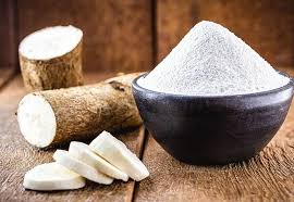 Finely ground cassava flour in a black bowl beside raw yuca root slices on a rustic wooden table.