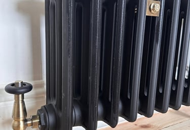 Vintage-style black cast iron radiator with brass thermostatic valves on a light wood floor.