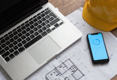 Top-down view of an engineer's desk with a high-end laptop, technical blueprints, and a smart device glowing with a sky blue interface.
