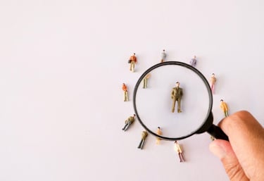 Hand holding a magnifying glass over a miniature person for recruitment and talent search.