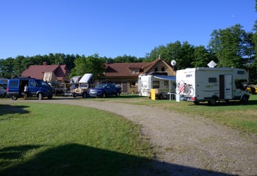 A sunny motorhome campsite with camper vans and RVs parked near a lodge in a grassy forest area.