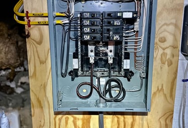 Open electrical breaker panel box with wired circuit breakers mounted on a wooden wall.