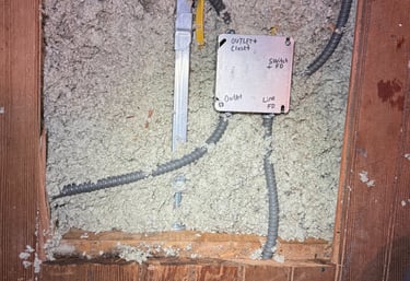 Electrical junction box with conduit wiring installed in blown-in attic insulation under wooden floorboards.