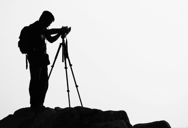 Silhouette of a travel photographer with a backpack using a tripod and camera on a rocky peak.