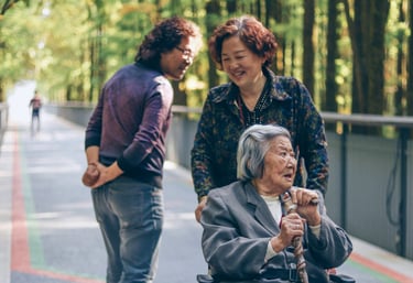A smiling woman pushes an older woman with grey hair in a wheelchair while a man walks past