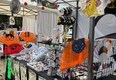 Puesto de venta artesanal en feria creativa con abanicos pintados a mano, stickers de anime y pop