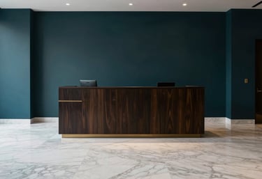 The minimalist reception area of a luxury consulting firm, with white marble floors and dark teal accent walls. A subtle gold logo is visible in the background. Global / Spanish-speaking.