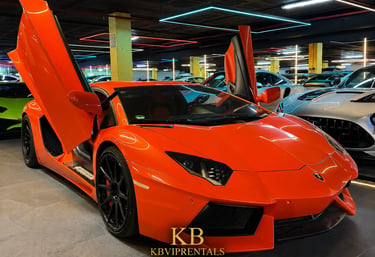 Bright orange Lamborghini Aventador with scissor doors open in a luxury car showroom.