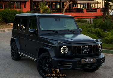 Matte black Mercedes-Benz G-Class luxury SUV rental parked in front of a tropical Marbella resort.