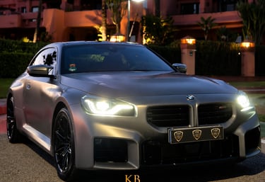 A matte grey BMW M2 sports car parked at sunset in front of a luxury resort in Marbella.