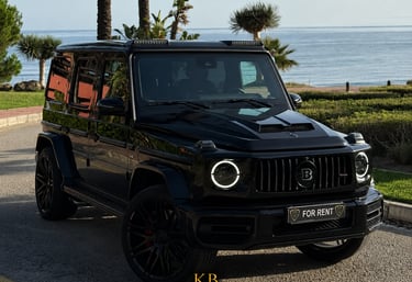 Luxury black Brabus G-Wagon rental parked by the sea in Marbella with palm trees.