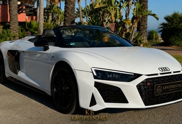 White Audi R8 Spyder convertible sports car parked outdoors in Marbella, Spain, surrounded by palm trees.