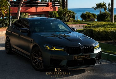 Matte black BMW M5 luxury rental car parked near a tropical Marbella beach resort.