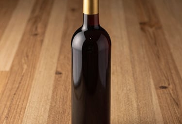 A professional product photograph of a dark burgundy wine bottle resting on an oak wood surface. Elegant lighting emphasizes the deep color of the glass and the gold foil on the neck.
