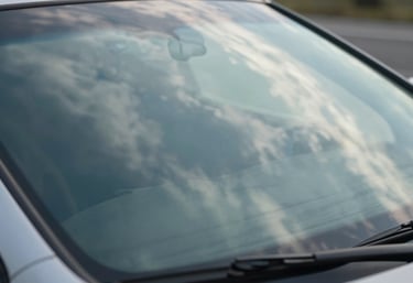 A macro photograph focusing on the surface of a polished, crystal-clear car windshield. Reflections of white clouds and a soft blue sky are visible in the glass, symbolizing perfect clarity and a job well done.