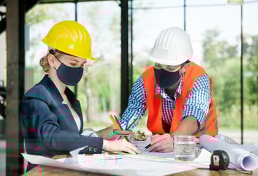 Two construction engineers wearing hard hats and face masks reviewing blueprints and architectural plans.